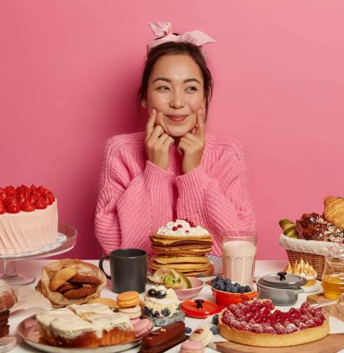 Photo of charming Korean woman keeps fingers on cheeks, surrounded with variery of sweet products, eats cakes, croissants and pancakes, tries to taste everything, wears headband and knitted jumper